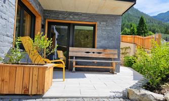 A cozy outdoor area with a wooden bench and yellow chairs. Surrounded by plants and a beautiful mountain landscape in the background.