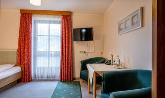 A simple hotel room with a bed, a table, and two armchairs. A window with curtains allows natural light in.