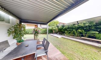 A cozy outdoor area with a table and chairs under a canopy. In the background, you can see a well-maintained garden with green plants.