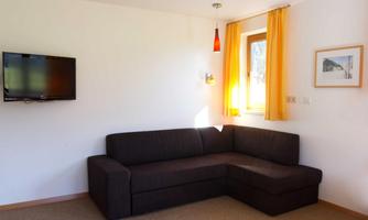 A bright living room with a chocolate brown sofa and a television on the wall. Large windows with yellow curtains allow for plenty of light.