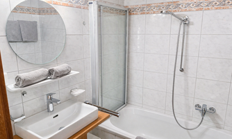 A modern bathroom with a bathtub, a shower, and a sink. The walls are tiled and there are towels on a shelf.