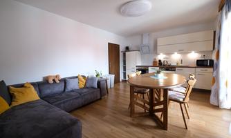 A modern living room with a gray sofa and a round dining table. The kitchen is bright and well-equipped, with warm wooden floors and natural light.