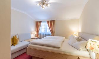 A bright bedroom with a large bed and a cozy sofa. The windows are decorated with curtains, and there is soft lighting.