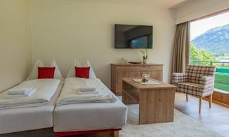 A modern room with two single beds and a television mounted on the wall. The room features a cozy seating area and a beautiful view of the mountains through the window.