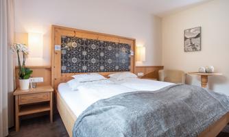 A cozy bedroom with a large bed and wooden furniture. The walls are bright and the decoration is simple, with a small table and a plant.