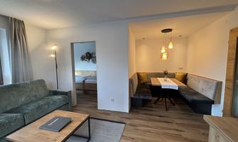 A modern living room with a green sofa and a wooden table. In the background, there is a dining area with a cozy bench and decorative lamps.