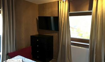 A cozy room with a large window and a TV on the wall. The room is furnished with curtains and a black wardrobe.