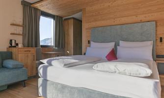 A cozy bedroom with a large double bed and soft colors. A window lets in daylight, and there is a small seating area with a table.