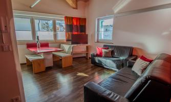 A modern and bright living room with a black sofa and a dining table. Large windows provide plenty of daylight.