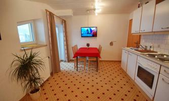 A cozy kitchen with a red table and chairs. A television hangs on the wall, and a small plant is in the corner.