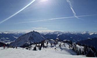 A wintry mountain landscape with lots of snow and clear visibility. Skiers can be seen on the slopes while the sun shines brightly in the sky.