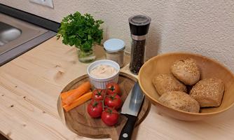 A wooden board with fresh ingredients, including carrots, tomatoes, and a dip. Next to the board, there are rolls in a bowl and spices.