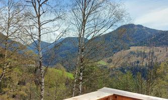 A beautiful view of green hills and mountains under a clear blue sky. In the foreground, there are birch trees and a wooden balustrade.