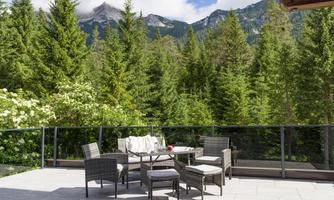A cozy terrace with a table and chairs made of rattan. In the background, green trees and mountains can be seen under a blue sky.