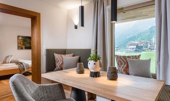 A modern living room with a wooden table and cozy chairs. Large windows offer a view of the surrounding landscape.
