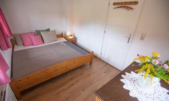 A cozy bedroom with a wooden bed, pillows, and colorful curtains. On a table, there is a vase with fresh flowers.