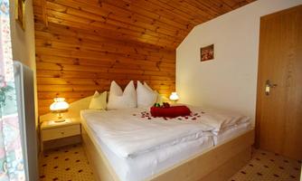 A cozy bedroom with a wooden ceiling and a large bed. On the bed are red rose petals and a decorative blanket.