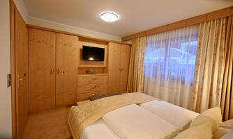 A cozy bedroom with a large bed and a wooden dresser. A television hangs on the wall and the windows are decorated with curtains.