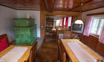 A cozy room with wooden tables and chairs. In the background, there is a green stove and there are shelves with dishes.