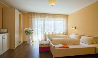 A bright bedroom with orange walls and a large window. There is a double bed, a chest of drawers, and a plant in a vase.