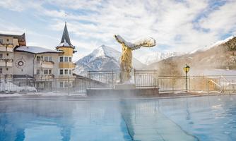 A heated outdoor pool surrounded by snow and mountains. In the background, there is a building with a tower and a statue.