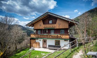 A traditional alpine house with a balcony and wooden cladding. In the background, mountains and a blue sky with clouds can be seen.
