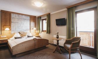 A cozy hotel room with a double bed and wooden decor. The large windows provide a view of a balcony and let in plenty of daylight.