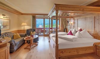 A stylish hotel room with a large four-poster bed and a cozy seating area. The windows offer a view of the surrounding nature.