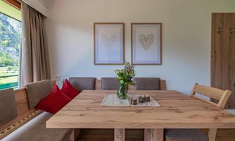 A bright living room with a large wooden table and comfortable benches. On the table, there is a vase with flowers and two pictures hang on the wall.