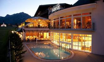 A modern hotel with large windows and an outdoor pool. The surroundings are mountainous and the evening sky is beautifully lit.
