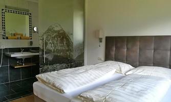 A modern hotel room with a double bed and a glass wall showcasing a mountain motif. On the wall, there is an elegant sink and a large mirror.