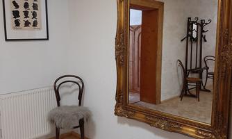 An elegant room with a large, golden mirror and a wooden chair draped with a fur throw. An artwork hangs on the wall, and a rug with an inscription lies on the floor.