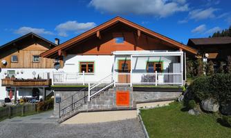 A modern house with an orange veranda and a large garden. In the background, traditional wooden houses can be seen under a blue sky with some clouds.