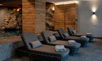 A tranquil wellness area with cozy loungers and a relaxing atmosphere. The walls are made of wood and stone, and in the background, a small pool can be seen.