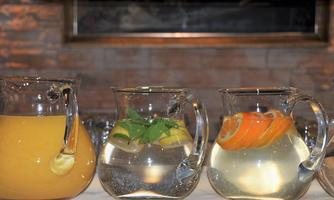 Three decorative jugs with beverages are on a table. The first contains a yellow juice, the second clear water with limes, and the third water with orange pieces.