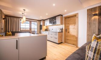 A modern living and kitchen area with wood details and warm lighting. Large windows provide daylight and a welcoming atmosphere.