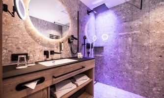 A modern bathroom with a large round mirror lamp and stylish fixtures. The walls are clad in elegant stone and there is an open shower with gentle lighting.