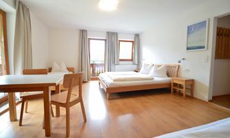A bright room with a double bed and a table with chairs. Large windows provide plenty of natural light.