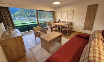 A modern living room with a red sofa, a wooden table, and a dining table. Large windows provide a view of the surrounding mountains and the green valley.