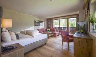 A modern room with wooden floors, a bed, and cozy seating. Large windows offer a view of the nature outside.