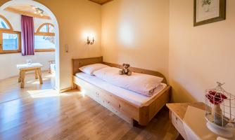 A cozy room with a wooden bed and a small table. Natural light streams through the windows, creating an inviting atmosphere.