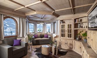 A cozy room with wooden paneling and large windows. The decor is simple and inviting, with plants and comfortable seating.