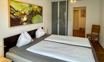 A modern bedroom with two beds and light wood flooring. A colorful painting hangs on the wall, and there is a chair in the corner.