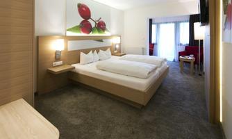 A modern hotel room with two single beds, bright colors, and a cozy atmosphere. Large windows let in daylight and offer a view of a relaxed seating area.