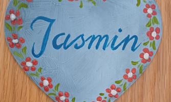 A heart-shaped sign in blue with the name "Jasmin". It is surrounded by small red and white flowers.