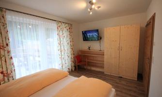 A cozy bedroom with two beds and a wooden dresser.  
Large windows with white curtains let in plenty of light.
