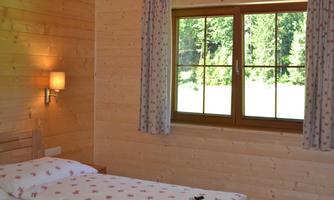 A cozy bedroom with wooden paneling and a window that overlooks a green landscape. A bed with a floral duvet cover and a lamp on the wall create an inviting atmosphere.