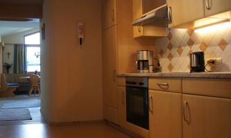 A modern kitchen with wooden cabinets and a stove. In the background, a cozy living area can be seen.