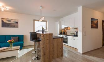 A modern kitchen with a wooden island and bar stools. The living area is bright and cheerful with a blue sofa and decor.