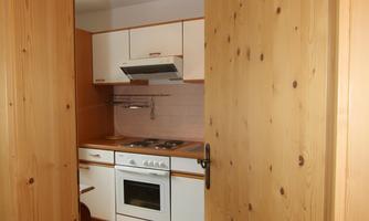 A bright kitchen with wooden shelves and modern appliances. The view leads through a wooden-paneled door.